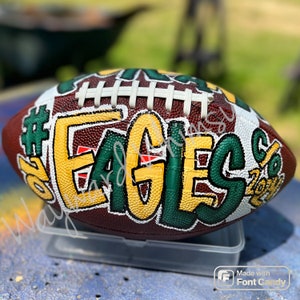 May include: A brown football with green, yellow, and white paint. The word "EAGLES" is written in large letters on the side of the football. The number "6" is painted on the side of the football. The words "2026" are painted on the side of the football.