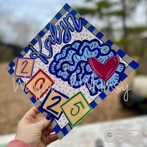 May include: A graduation cap decorated with a blue brain, a pink heart, and colorful blocks with the numbers 2021. The name "Kalyan" is written in blue cursive on the cap.