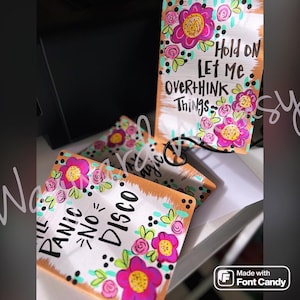 May include: Two painted wooden signs with floral designs and inspirational quotes. The first sign says "Hold on Let Me Overthink Things" and the second sign says "Ill Panic No - Disco".
