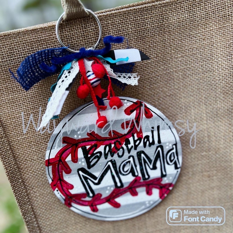 Może przedstawiać: Biało-szary okrąg z czerwonymi szwami baseballowymi i napisem "Baseball Mama" czarnymi literami. Okrąg jest przymocowany do breloka z niebieskimi, białymi i czerwonymi ozdobami.