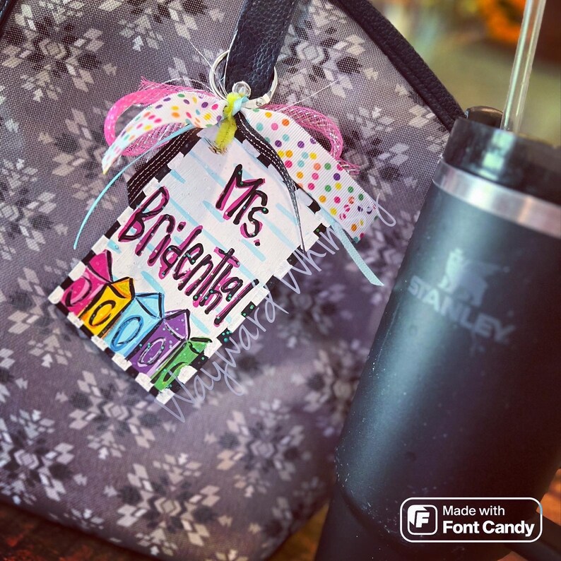 May include: A colorful teacher name tag with the text "Mrs. Bridenthal" and a colorful design of presents. The tag is attached to a black bag with a white and gray pattern.