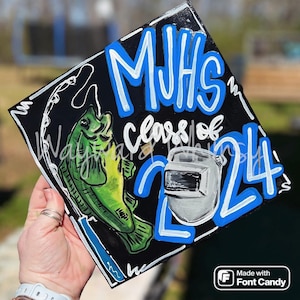 May include: A black graduation cap decorated with blue and white paint. The cap features the text "MJHS Class of 2024" and a green fish with a fishing line. A welding mask is also painted on the cap.