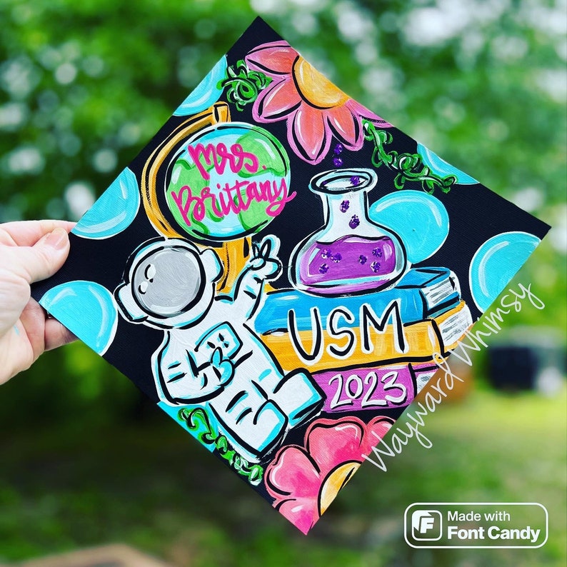 May include: A black graduation cap decorated with a colorful astronaut, a globe, a beaker, books, flowers, and the text "Mrs. Brittany", "USM", "2023", and "Wayward Whimsy".