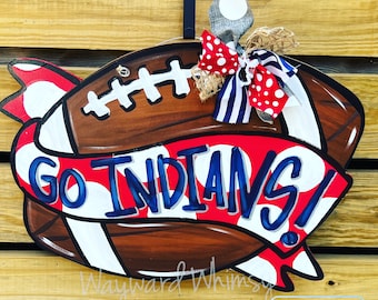 Peut inclure: Un porte-manteau de football en bois avec un ruban rouge, blanc et bleu qui dit "Go Indians!" Le ballon de football est brun avec des lacets blancs et des rayures blanches. Le ruban a des pois blancs sur un fond rouge.