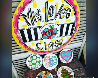 May include: A set of six round wooden signs with various holiday themes. The signs are painted in bright colors and feature festive designs, including a turkey, a Christmas tree, a shamrock, a heart, a ghost, and an apple. The signs are perfect for decorating a classroom or home for the holidays.