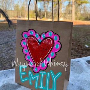 May include: A brown paper gift bag with a hand-painted design. The design features a red heart with a pink and teal border. The name "Emily" is written in teal below the heart, and the words "Wayward Whimsy" are above the heart.
