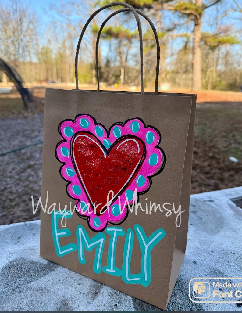 May include: A brown paper gift bag with a hand-painted design. The design features a red heart with a pink and teal border. The name "Emily" is written in teal below the heart, and the words "Wayward Whimsy" are above the heart.