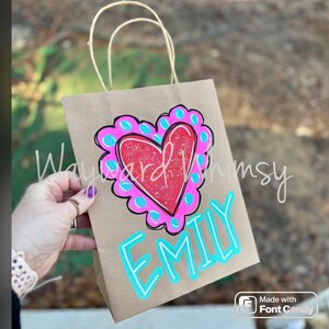 May include: A brown paper gift bag with a hand-painted heart design. The heart is red with a pink and blue border. The name "EMILY" is written in blue. The bag has brown paper handles.