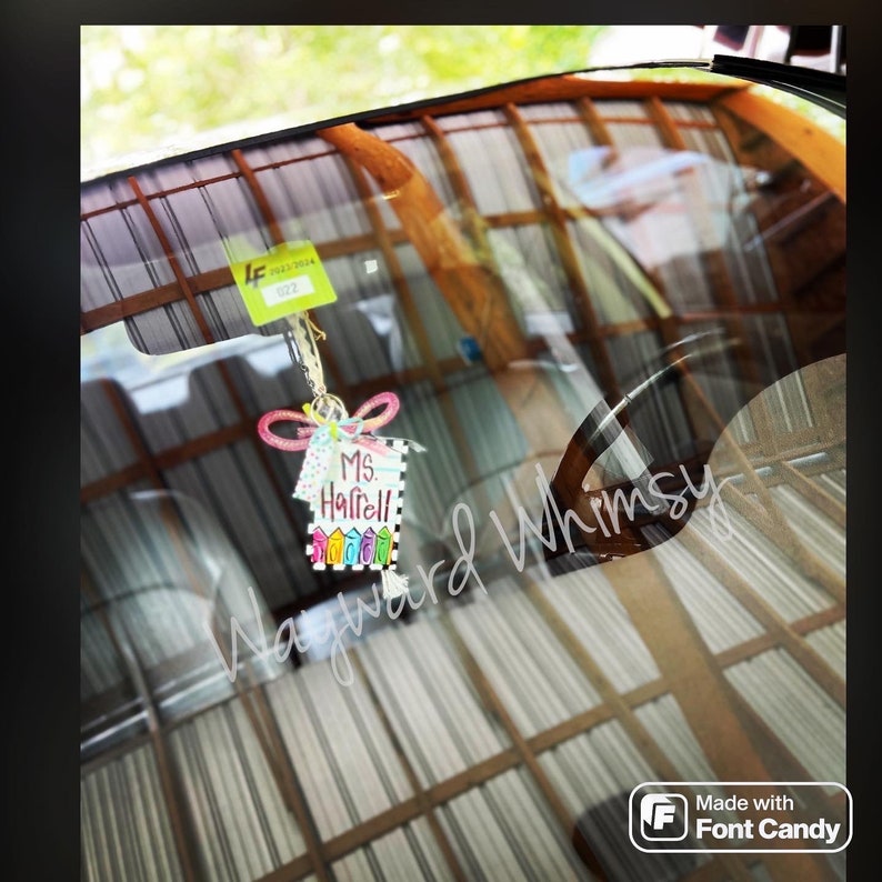 May include: A colorful car air freshener with the text "Ms. Harrell" and a rainbow design. The air freshener is hanging from the rearview mirror of a car.