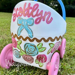 May include: A white plastic toy stroller with four pink wheels and a blue handle. The front is hand-painted with the name Scottlyn in pink, a blue bow, a gold wavy line, and a floral pattern featuring blue, pink, and green painted roses.