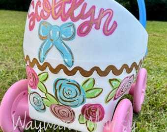 May include: A white plastic toy stroller with four pink wheels and a blue handle. The front is hand-painted with the name Scottlyn in pink, a blue bow, a gold wavy line, and a floral pattern featuring blue, pink, and green painted roses.
