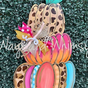 May include: A stacked pumpkin-shaped wooden door hanger with a teal stem and a decorative bow. The pumpkins are painted in various colors, including tan with brown spots, pink, orange, and blue. The bow features pink, gold, and leopard print ribbons.