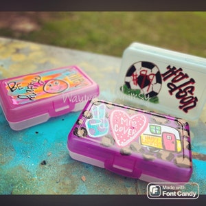 May include: Three colorful pencil cases with different designs. One case has a pink background with the words "Be Happy" and a smiley face. Another case has a purple background with a peace sign, a heart with the words "Mrs. Cover", and a camper van. The third case has a white background with a soccer ball and the name "Hudson".