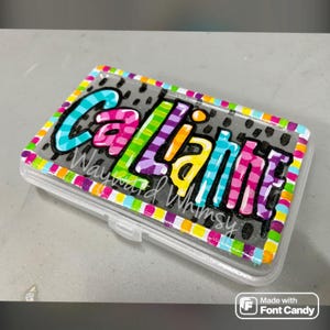 May include: A clear plastic case with a colorful design. The lid features the name "Callie" in vibrant, multi-colored lettering, with "Wayward Whimsy" below. The border is decorated with a rainbow pattern.