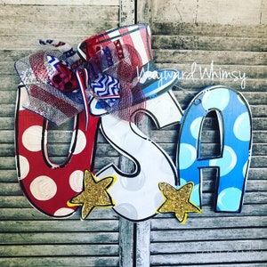 May include: A wooden USA door hanger with red, white, and blue polka dots and stars. The letters are painted white with gray polka dots. A red, white, and blue bow and a red, white, and blue top hat are attached to the top of the hanger. The text "Wayward Whimsy" is visible in the top right corner of the image.