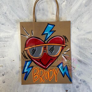 May include: A brown paper gift bag with a red heart wearing sunglasses and the name "BRODY" in orange. The heart has a black outline and orange sunglasses. Blue lightning bolts are on the bag.