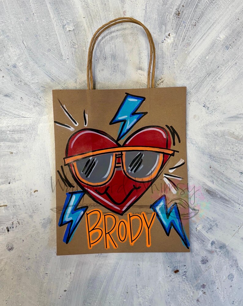 May include: A brown paper gift bag with a red heart wearing sunglasses and the name "BRODY" in orange. The heart has a black outline and orange sunglasses. Blue lightning bolts are on the bag.