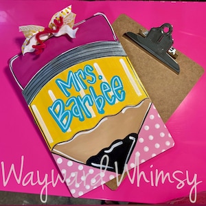 May include: A clipboard shaped like a pencil with a pink eraser top, a yellow body, and a black lead tip. The text Mrs. Barbee is written in blue script. A decorative bow with yellow and white patterns sits at the top. The clipboard has a metal clip.