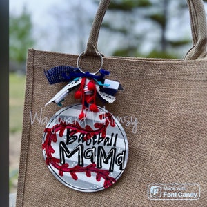 Może przedstawiać: Biało-szary brelok w kształcie piłki baseballowej z czerwonymi szwami i napisem "Baseball Mama" w kolorze czarnym. Brelok jest przymocowany do brązowej torby z juty.
