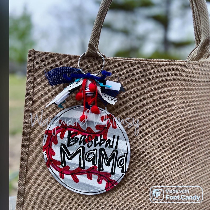 Może przedstawiać: Biało-szary brelok w kształcie piłki baseballowej z czerwonymi szwami i napisem "Baseball Mama" w kolorze czarnym. Brelok jest przymocowany do brązowej torby z juty.