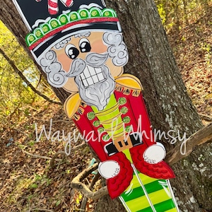 May include: A wooden Christmas nutcracker decoration. The nutcracker has a black hat with candy canes, a white mustache and beard, and a red and gold uniform. The legs are green and yellow striped. The text "Wayward Whimsy" is written on the decoration.