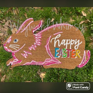 May include: A brown coir doormat shaped like a bunny rabbit with pink outlines. The words "Happy Easter" are written in white with the word "Easter" in rainbow colors.