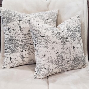 May include: Two white throw pillows with a black and white world map print. The pillows feature a compass and airplanes on the map. The text "THE WORLD" is visible on one of the pillows.