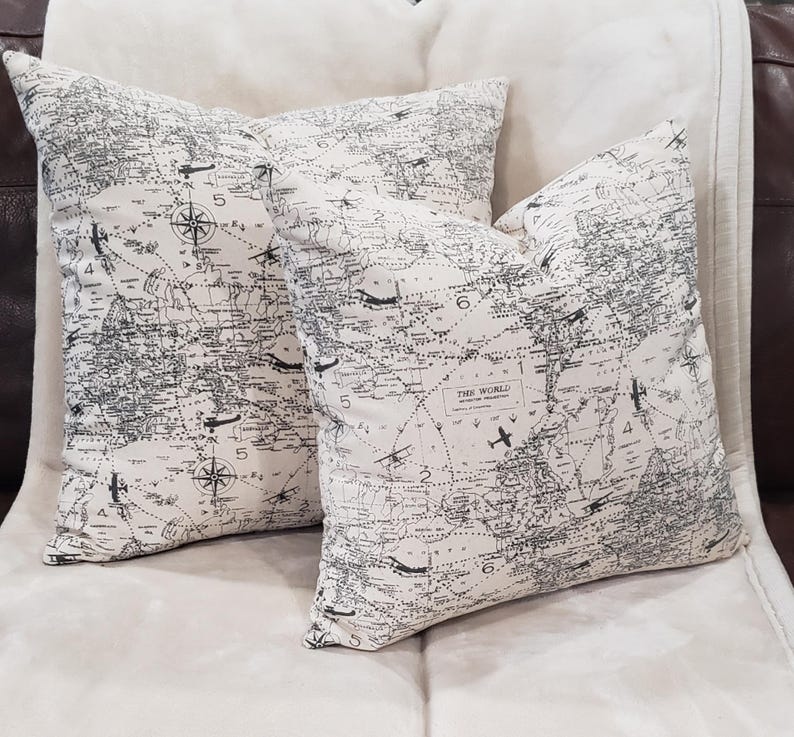 May include: Two white throw pillows with a black and white world map print. The pillows feature a compass and airplanes on the map. The text "THE WORLD" is visible on one of the pillows.