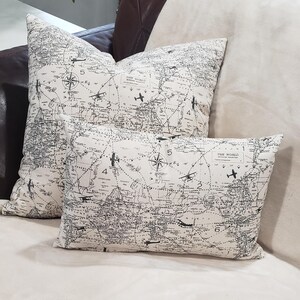 May include: Two decorative throw pillows with a vintage world map design featuring airplanes and compass roses. The pillows are on a cream-colored couch.
