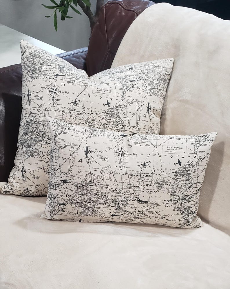May include: Two decorative throw pillows with a vintage world map design featuring airplanes and compass roses. The pillows are on a cream-colored couch.