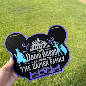 May include: A black, mouse-ear shaped sign with a purple border featuring light blue silhouettes of characters, a mansion, and a gate. The text reads: THIS DOOM BUGGY BELONGS TO THE ZAPIEN FAMILY. The sign includes two horizontal slots at the top.
