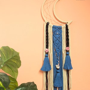 May include: A macrame wall hanging in a crescent moon shape. The design features cream, black, and blue woven elements, with blue tassels and small pink floral accents. The Boho Crafts logo is visible in the upper right corner.