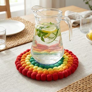 Può includere: Una caraffa di vetro trasparente piena d'acqua, fette di limone e menta poggia su un sottopentola di feltro colorato. Il sottopentola è un arcobaleno di palline di feltro rosse, arancioni, gialle, verdi e blu.