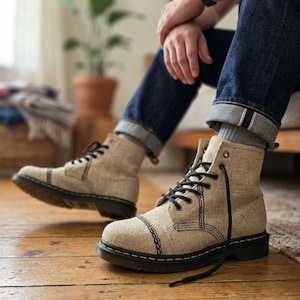 May include: A pair of beige canvas lace-up boots with black stitching details, black laces, and thick black rubber soles. The boots feature a reinforced toe cap design with visible black thread accents and silver-toned metal eyelets for the laces.