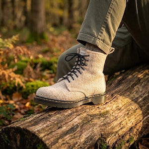 May include: A beige ankle boot with a textured fabric upper, black laces, and a dark sole with yellow stitching. A small white tag at the top of the tongue features the word HEMP in black text. The boot has a rounded toe and a pull tab at the heel.