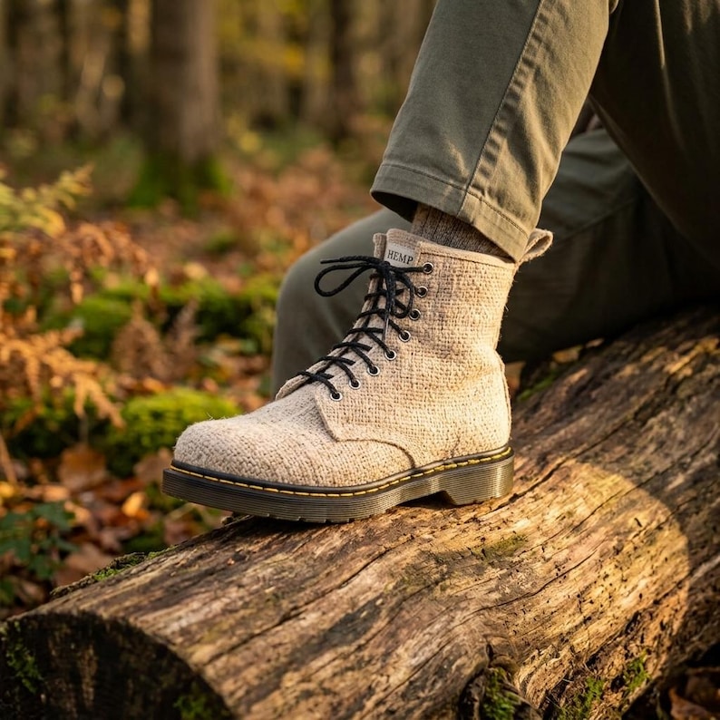 May include: A beige ankle boot with a textured fabric upper, black laces, and a dark sole with yellow stitching. A small white tag at the top of the tongue features the word HEMP in black text. The boot has a rounded toe and a pull tab at the heel.