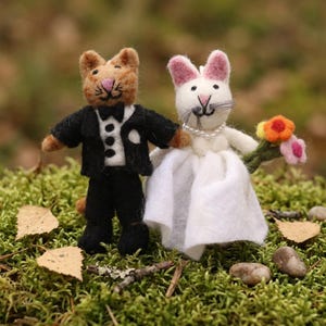 Op de afbeelding: Twee vilten kattenbeeldjes, een bruid en bruidegom. De bruidegom is bruin, draagt een zwart smoking en een vlinderdas. De bruid is wit, in een witte jurk en parelketting, met een klein boeket. Ze staan op groen mos.