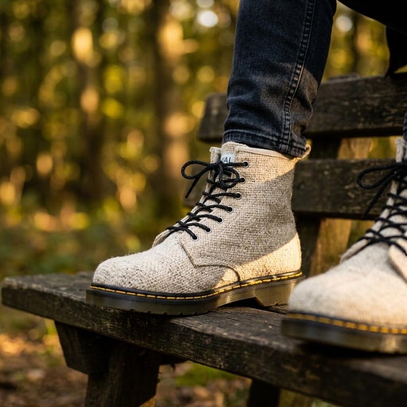 May include: A pair of beige, textured fabric ankle boots with black laces and a thick, dark brown sole featuring yellow stitching. The boots have a small white label on the tongue with the text "VAL" visible on the upper portion of the boot.