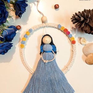 May include: A handmade Boho Crafts wall hanging featuring a doll with a blue dress and black hair, suspended within a macrame ring. The ring is decorated with colorful floral accents. The background includes blue flowers and decorative elements.