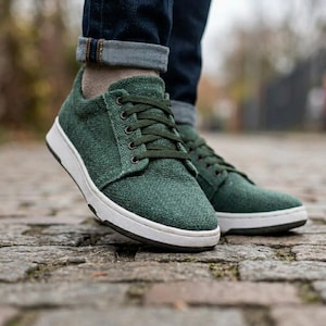 May include: Green sneakers with white soles and dark green laces. The shoes have a textured fabric upper and are worn with blue jeans. The sneakers are designed for casual wear and outdoor activities.