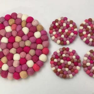May include: A collection of felt ball coasters and a matching trivet. The trivet and coasters are made of small, colourful felt balls in shades of pink, white, and tan. The trivet is round, and the coasters are also round.