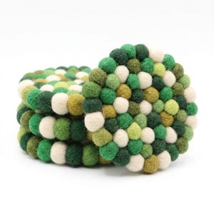 May include: A circular coaster constructed from numerous small, densely packed felt balls in various shades of green, olive, and cream. The individual spheres are tightly joined to form a thick, textured surface with a uniform, rounded edge.