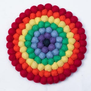 May include: A circular, multi-coloured felt trivet in a rainbow pattern. The trivet is made of small, round felt balls in shades of red, orange, yellow, green, blue, and purple. The trivet is approximately 20 cm in diameter.