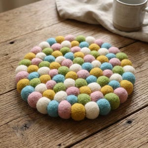 Puede incluir: Un salvamanteles redondo hecho de bolas de fieltro de colores en tonos pastel de rosa, azul, verde, amarillo y blanco. El salvamanteles está colocado sobre una superficie de madera, con una taza y un paño de lino en el fondo. El salvamanteles tiene unos 20 cm de diámetro.