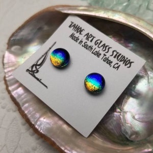 Puede incluir: Dos pendientes de bot&oacute;n redondos de vidrio dicr&oacute;ico con una vibrante mezcla de colores, incluyendo azul, verde y dorado. Los pendientes se muestran en una tarjeta blanca con el texto "TAHOE ART GLASS STUDIOS Made in South Lake Tahoe, CA".
