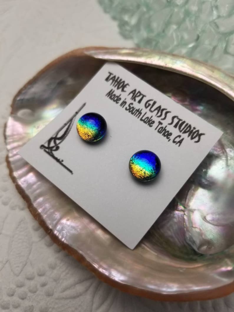 Puede incluir: Dos pendientes de bot&oacute;n redondos de vidrio dicr&oacute;ico con una vibrante mezcla de colores, incluyendo azul, verde y dorado. Los pendientes se muestran en una tarjeta blanca con el texto "TAHOE ART GLASS STUDIOS Made in South Lake Tahoe, CA".