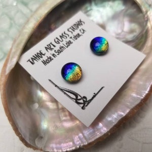 Puede incluir: Pendientes de bot&oacute;n de vidrio dicr&oacute;ico en azul, verde y dorado, presentados sobre una tarjeta blanca dentro de una concha iridiscente. La tarjeta dice "TAHOE ART GLASS STUDIOS Made in South Lake Tahoe, CA."