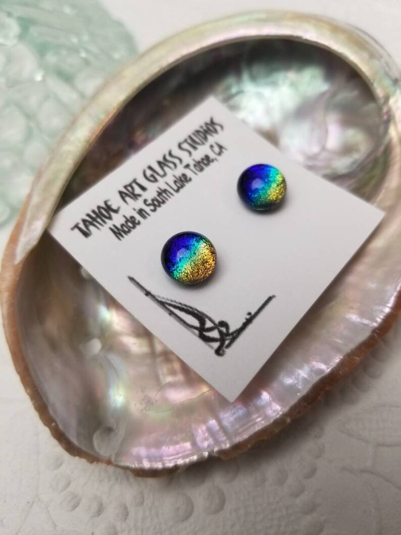 Puede incluir: Pendientes de bot&oacute;n de vidrio dicr&oacute;ico en azul, verde y dorado, presentados sobre una tarjeta blanca dentro de una concha iridiscente. La tarjeta dice "TAHOE ART GLASS STUDIOS Made in South Lake Tahoe, CA."