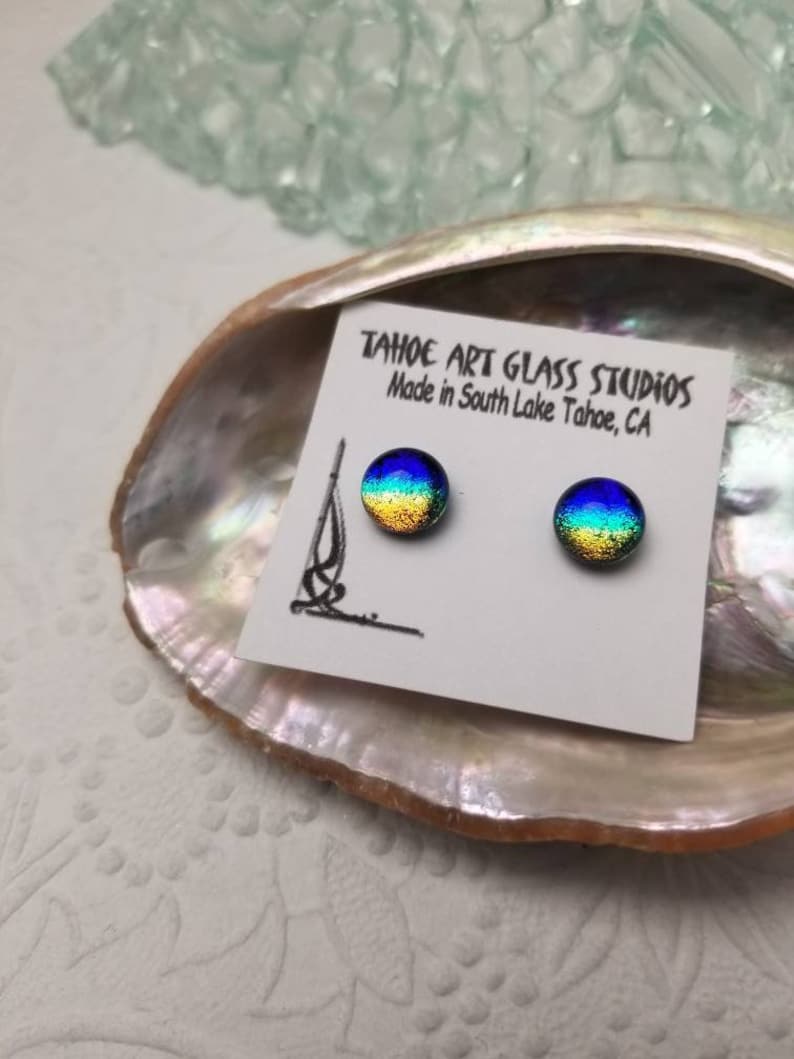 Puede incluir: Un par de pendientes redondos de cristal dicroico con un patr&oacute;n de arco&iacute;ris, expuestos en una tarjeta blanca dentro de una concha. La tarjeta dice "TAHOE ART GLASS STUDIOS Made in South Lake Tahoe, CA."