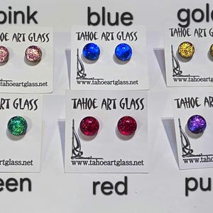 May include: Six pairs of round stud earrings in pink, blue, gold, green, red, and purple, each mounted on a white card labeled TAHOE ART GLASS with the website www.tahoeartglass.net. The glass earrings feature a textured, glittery appearance.